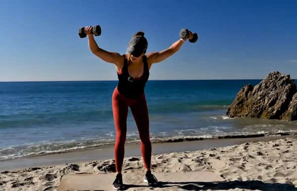 A person in workout clothes performs a bent-over dumbbell exercise on a sandy beach, facing the ocean, with arms raised and weights in each hand.