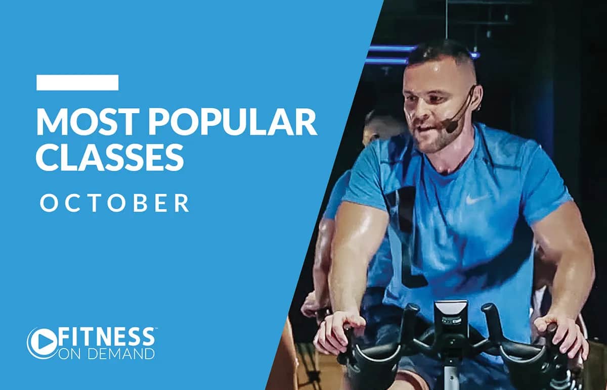 A fitness instructor wearing a blue shirt leads a cycling class with a headset microphone. Text on a blue background reads Most Popular Classes October and Fitness On Demand with a circular logo.