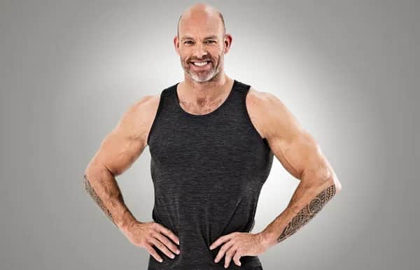 Bald, muscular man with a beard and tattoos, wearing a dark sleeveless shirt, smiles confidently with hands on hips against a plain grey background.
