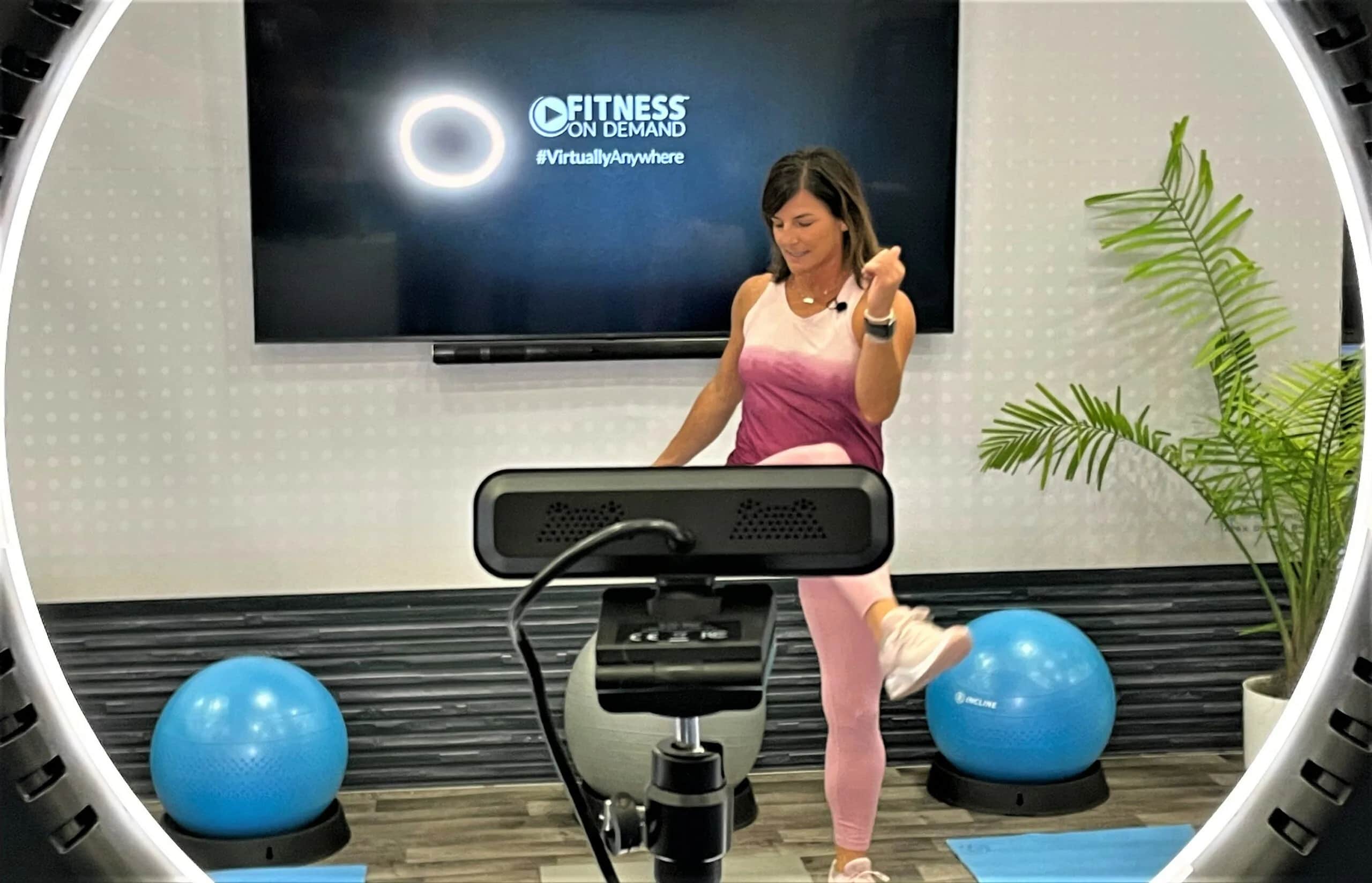 A woman in workout clothes exercises in front of a camera and ring light. Behind her, a screen displays “Fitness on Demand #VirtuallyAnywhere.” Two blue exercise balls and a plant are in the room.