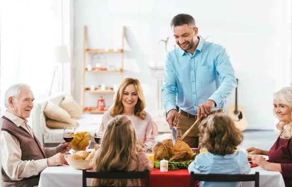 A family of six gathers around a dining table, smiling as a man carves a roast chicken. Adults and children are seated, enjoying food and drinks in a bright, cozy living room.