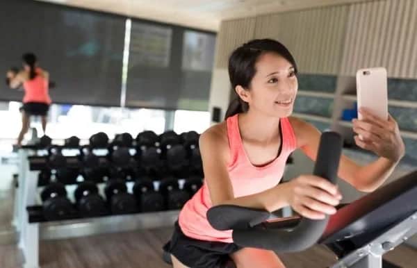A woman in a pink tank top is smiling and taking a selfie while using an exercise bike in a gym with dumbbells and mirrors in the background.