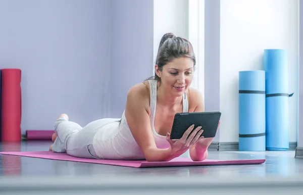 A woman in workout clothes lies on a yoga mat indoors, looking at a tablet. Yoga mats are rolled up in the background, suggesting she is in a fitness or yoga studio.