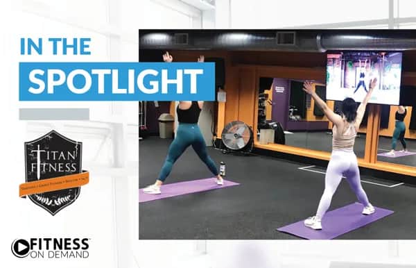 Two women on yoga mats in a gym stretch with arms raised, following a fitness class on a large screen. In the Spotlight and gym logos Titan Fitness and Fitness On Demand appear on the left.