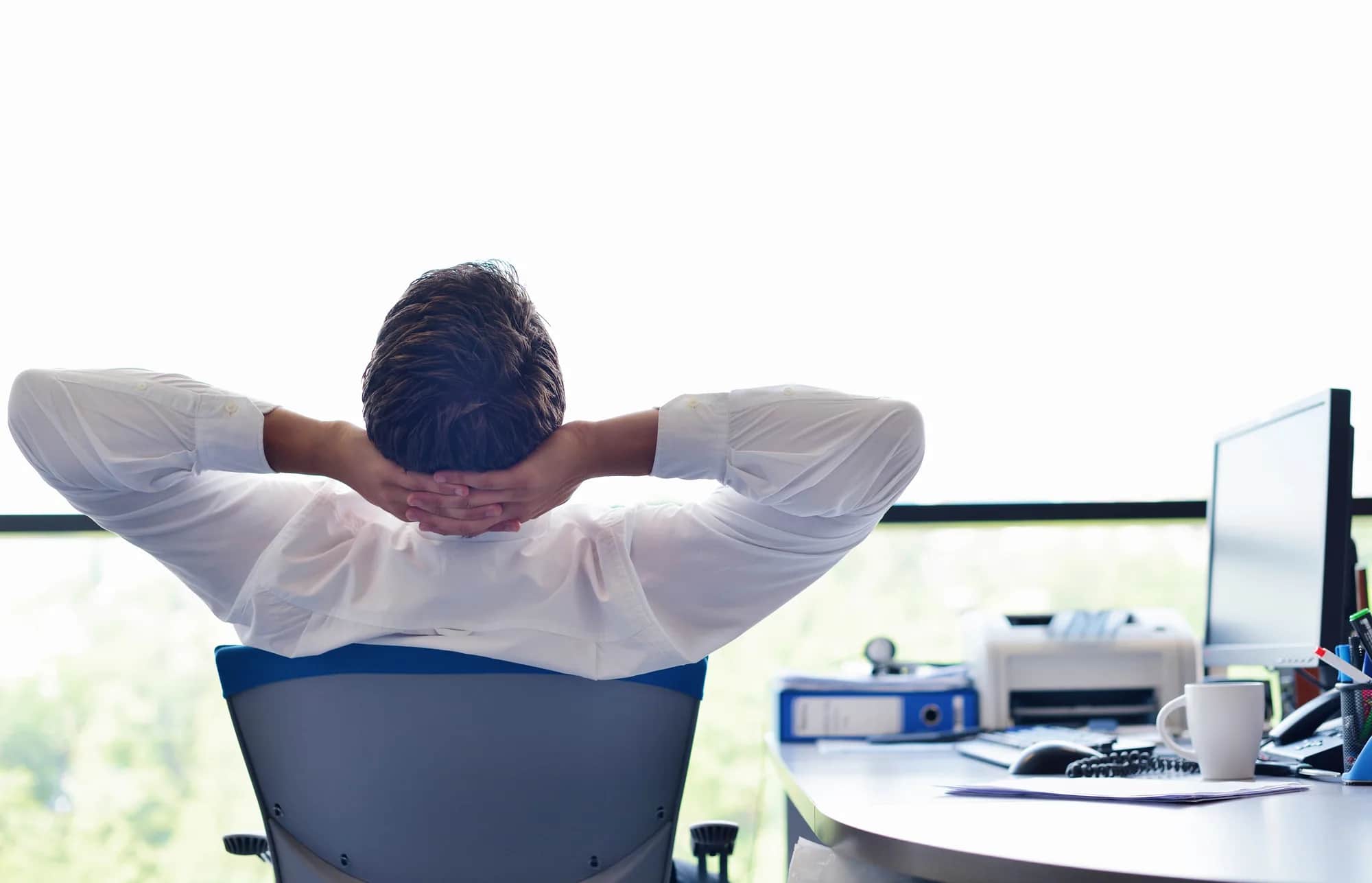 A person in a white shirt sits at a desk with hands behind their head, looking out a bright window. Office items, a computer, and a coffee cup are on the desk, suggesting a relaxed work environment.