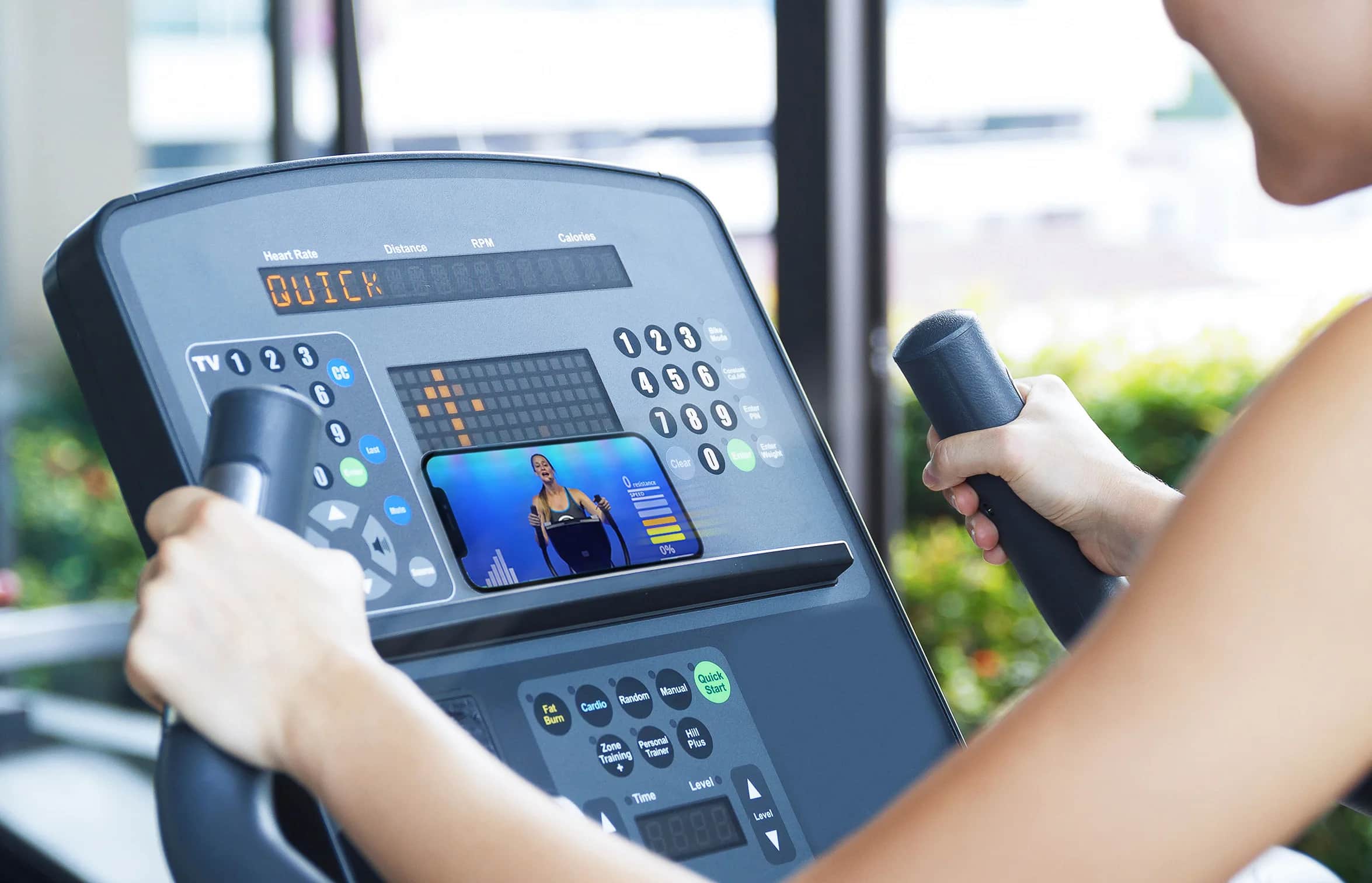 A person uses an elliptical machine, holding the handlebars, while watching a fitness video on a smartphone placed above the machines display panel. Large windows reveal greenery outside.