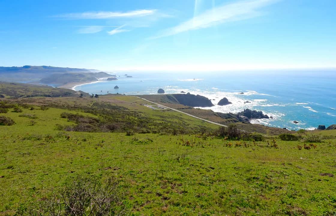 A scenic coastal landscape with rolling green hills in the foreground, a winding road, rocky cliffs, and the blue ocean stretching to the horizon under a bright sky.