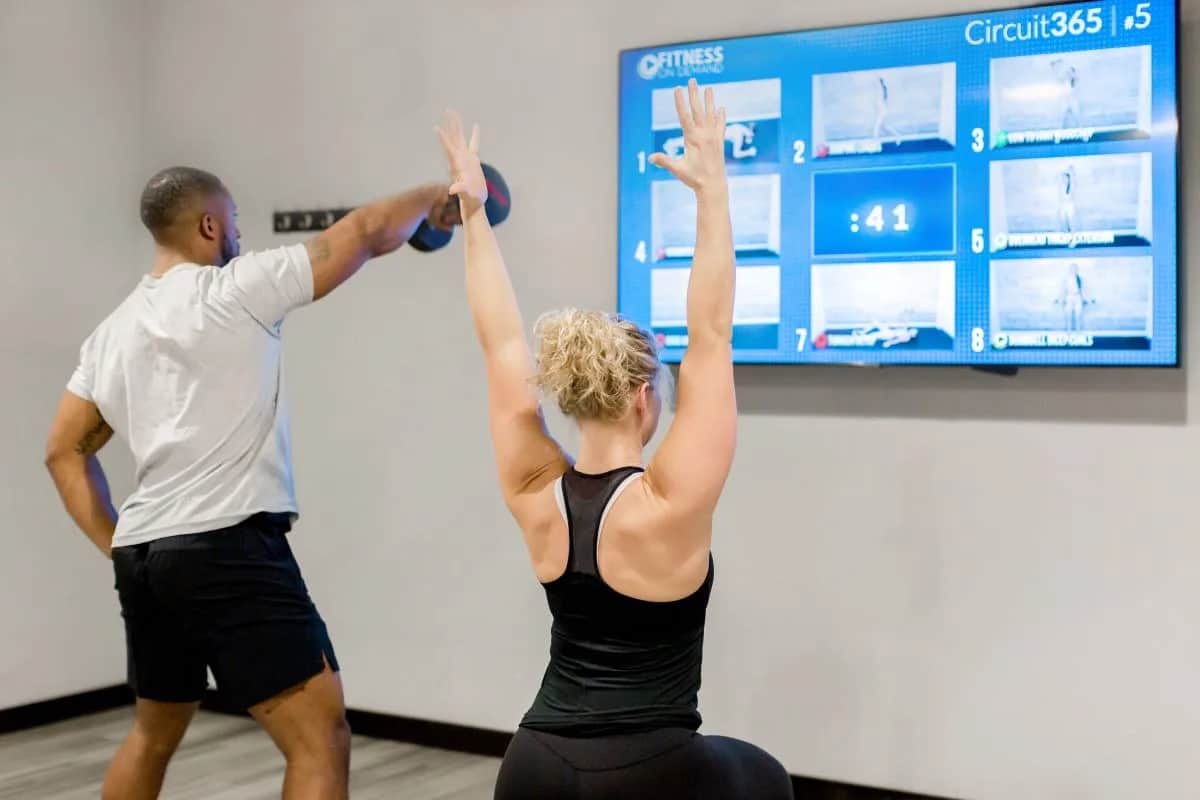 Two people exercise in a gym, following a workout displayed on a wall-mounted screen. One lifts a dumbbell, and the other raises her arms overhead; both are facing the screen showing fitness routines and a timer.