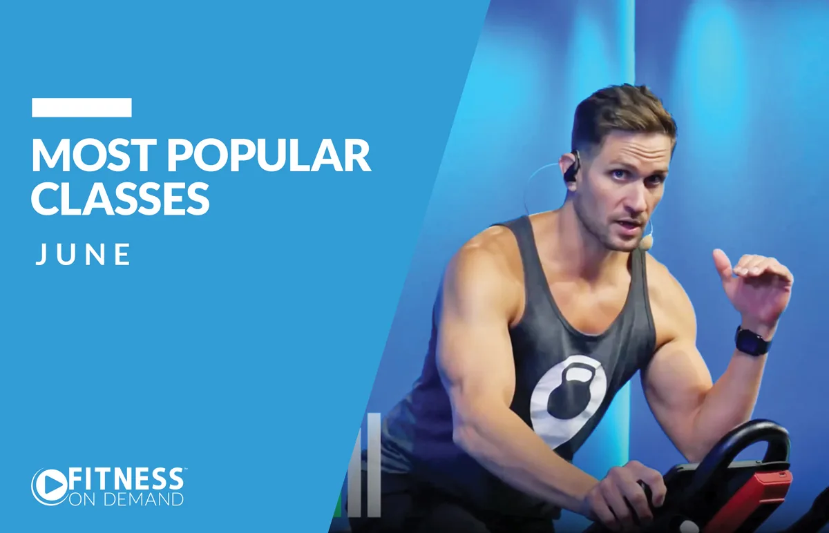 A fit instructor wearing a headset and tank top leads an indoor cycling class on a stationary bike. The left side features text: Most Popular Classes June with the Fitness On Demand logo below on a blue background.