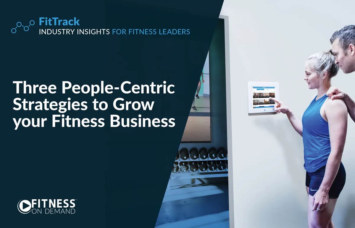 A woman in athletic wear uses a wall-mounted touchscreen in a gym, guided by a male trainer. Text on the left reads, “Three People-Centric Strategies to Grow your Fitness Business.” Logos for FitTrack and FITNESS On Demand appear nearby.