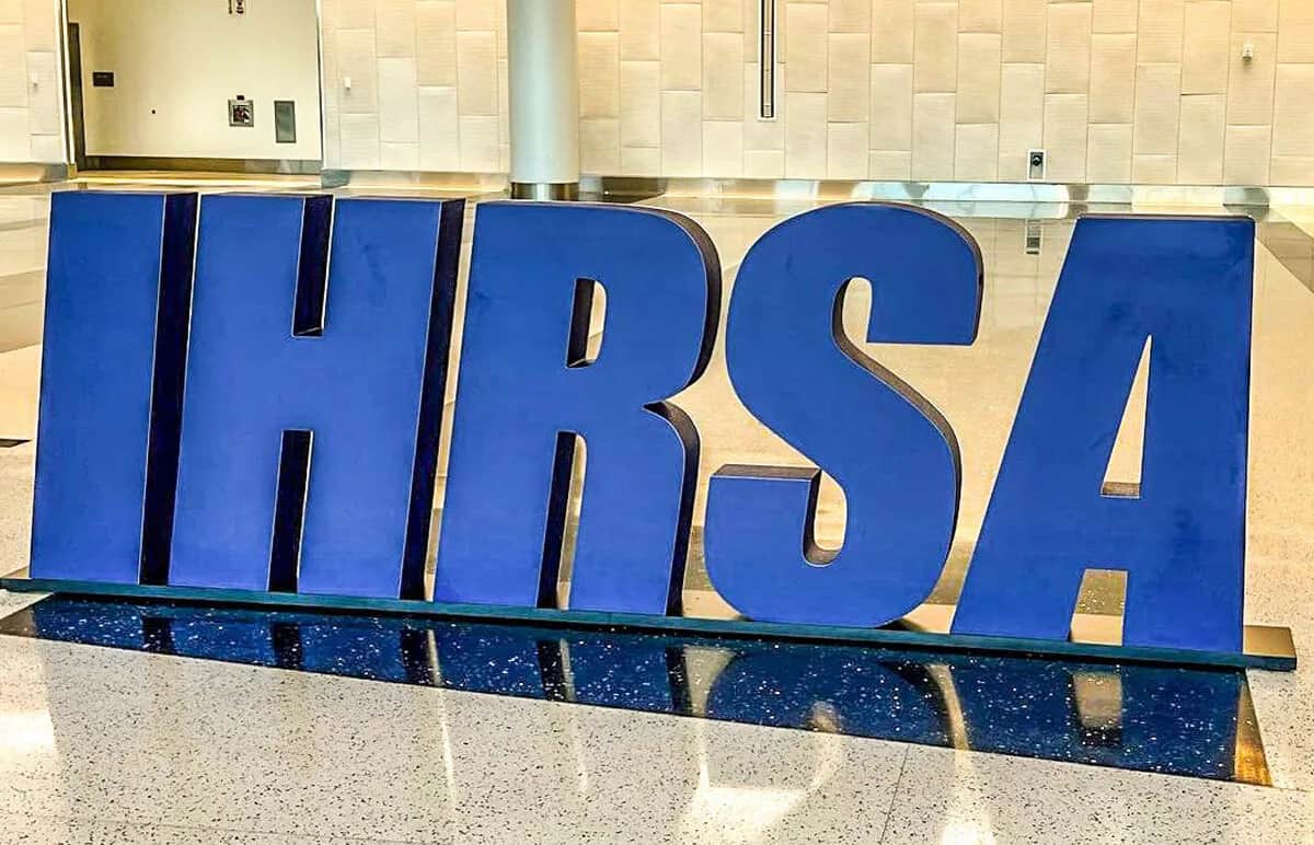 Large blue letters spelling IHRSA are displayed indoors on a shiny floor with a light-colored, textured wall in the background.