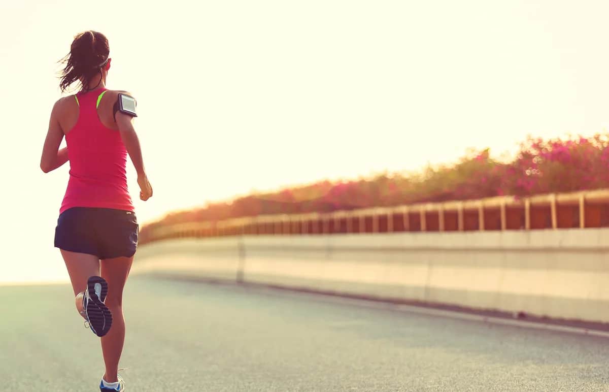 A woman in athletic wear runs on an empty road beside a concrete barrier, with sunlight and greenery in the background. She has a phone armband and is captured from behind.