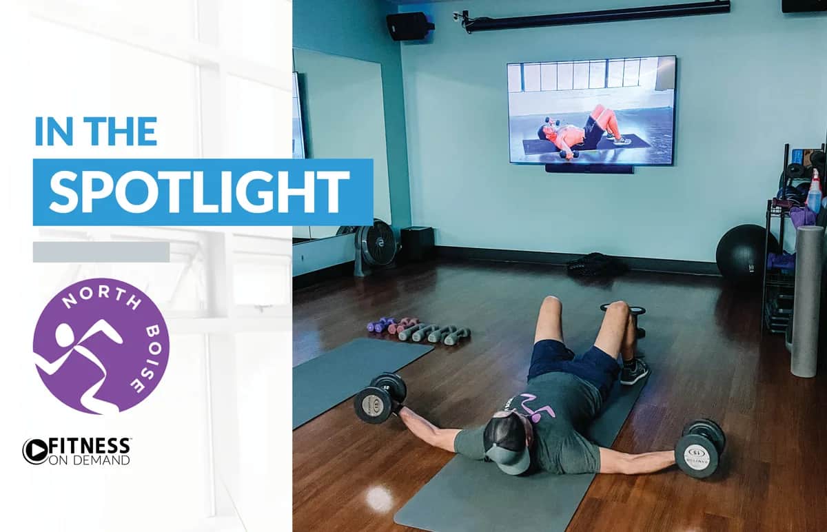 A person lies on a mat in a gym, lifting dumbbells. A fitness video is playing on a large screen in front. The text reads “IN THE SPOTLIGHT” with the North Boise logo and “Fitness On Demand” on the left.
