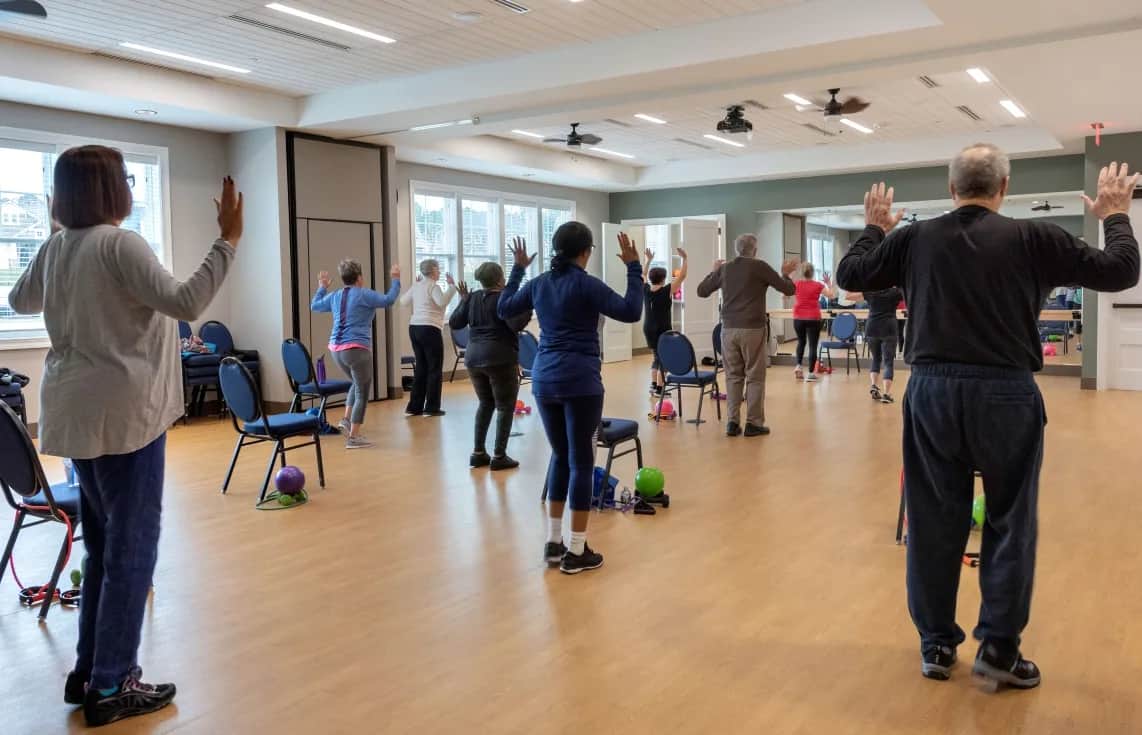 A group of older adults participate in a fitness class in a bright room, standing behind chairs with their arms raised. Exercise equipment, such as balls and bands, is placed nearby.