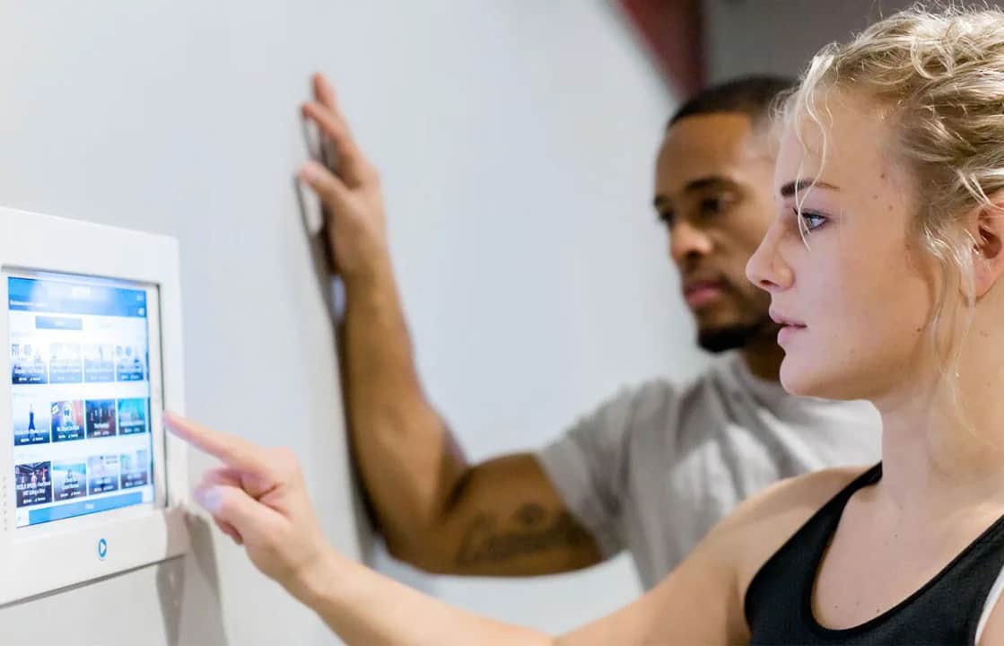A woman touches a wall-mounted touchscreen displaying various options, while a man stands beside her, watching. Both appear focused and are dressed in athletic clothing.