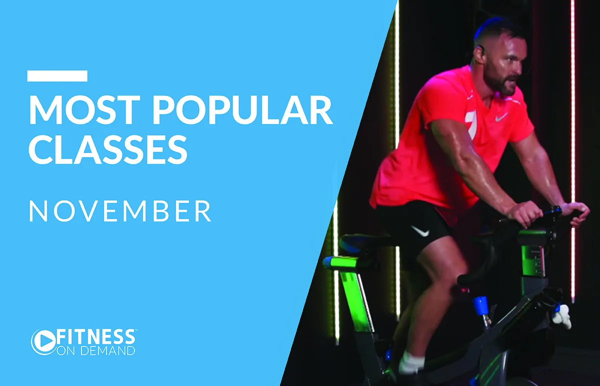 A man in a bright pink shirt rides a stationary bike indoors. Text on a blue background reads: “Most Popular Classes November.” The Fitness On Demand logo appears at the bottom left.