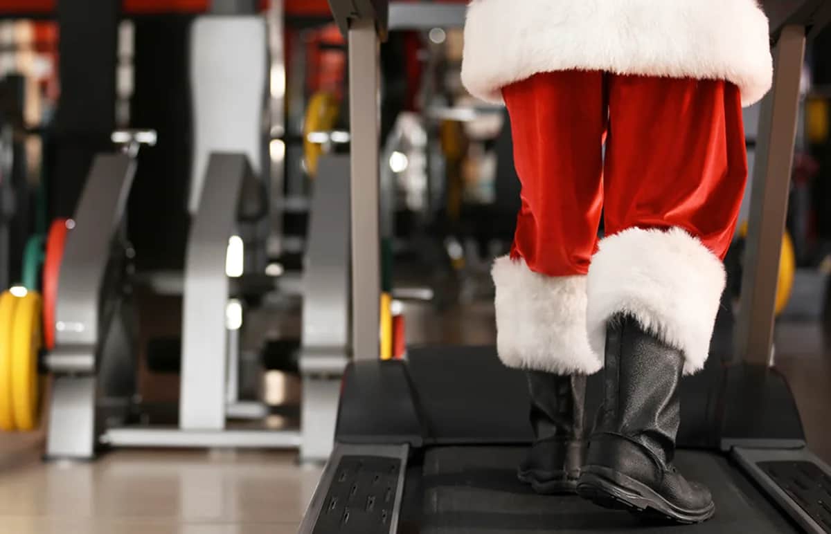 Person dressed in a Santa Claus costume, visible from the waist down, walking on a treadmill in a gym with exercise equipment in the background.