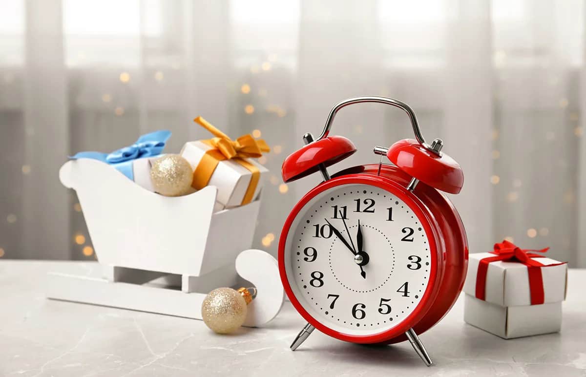A red alarm clock shows the time as 11:55. Beside it are a white sleigh with gold and silver ornaments and small wrapped gifts with bows. The background is softly lit with white curtains and blurred lights.