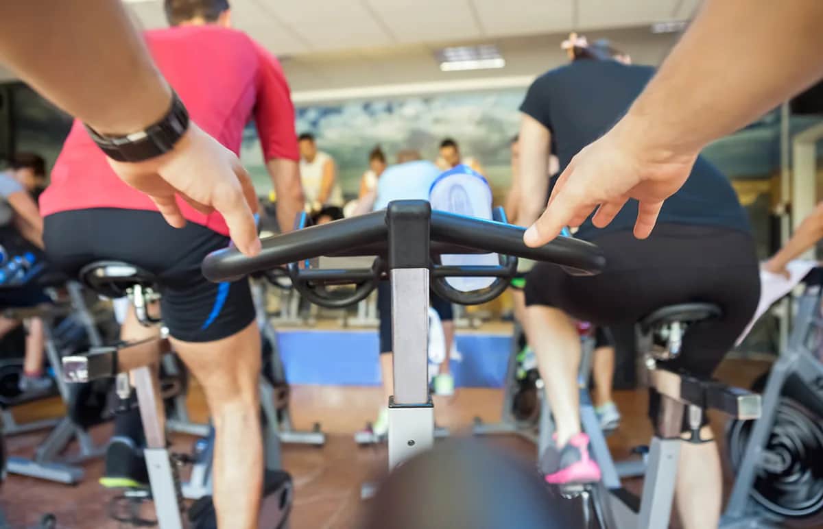 Close-up view from behind of people participating in a spin class, riding stationary bikes indoors; focus is on the handlebars and backs of cyclists wearing athletic clothing.