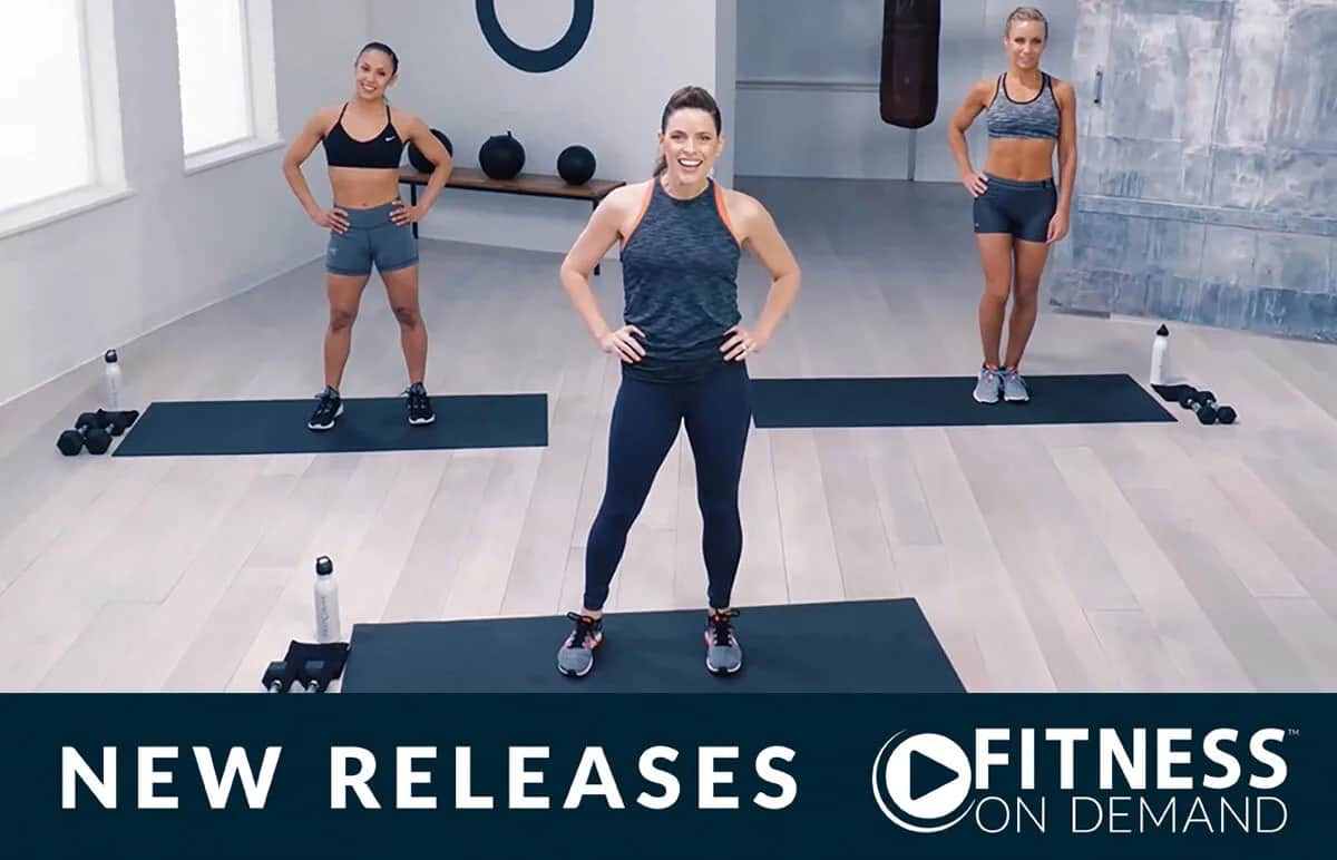 Three women in fitness attire stand on exercise mats in a bright studio, smiling and posing for a workout. The text “NEW RELEASES” and “FITNESS ON DEMAND” appears at the bottom of the image.