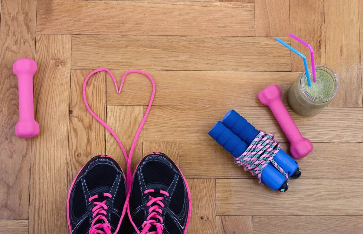 Overhead view of black sneakers with pink laces, a pink shoelace shaped like a heart, two pink dumbbells, a blue jump rope, and a green smoothie in a jar with blue and pink straws on a wooden floor.