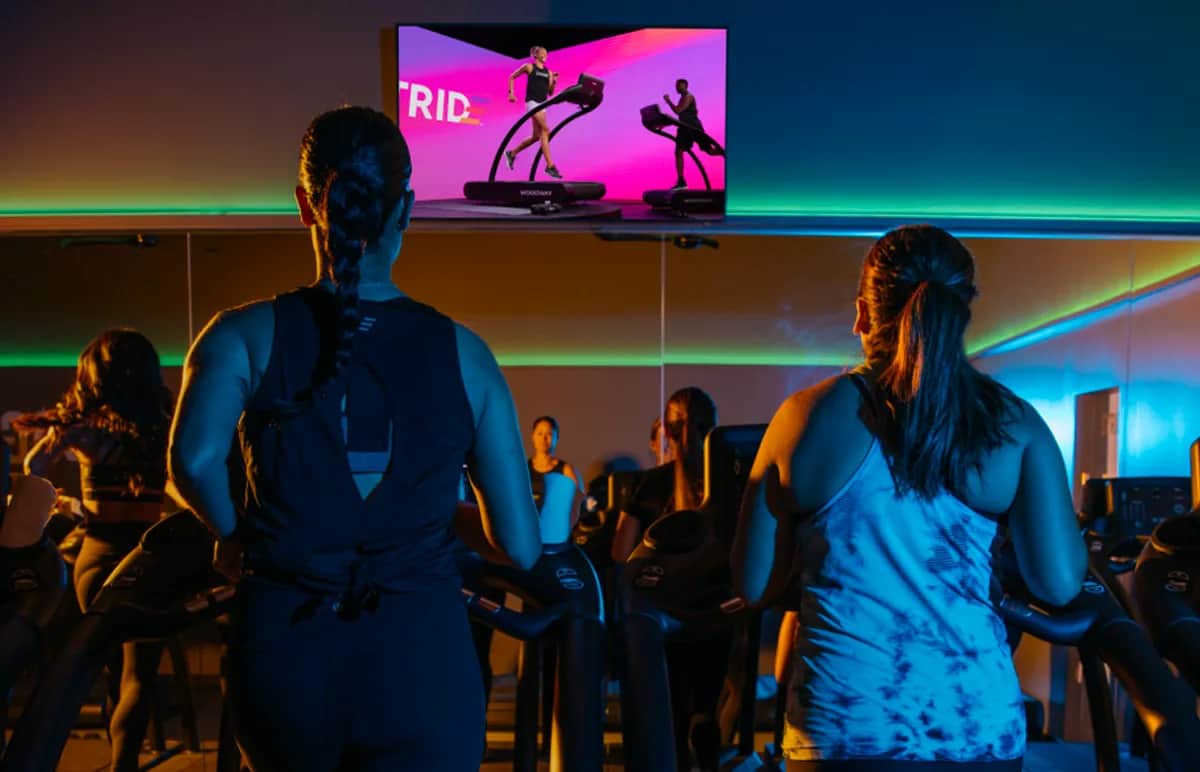 Two women exercise on treadmills in a gym, watching a fitness class on a large screen in front of them. The room is dimly lit with colorful neon lights and a mirrored wall reflecting the scene.
