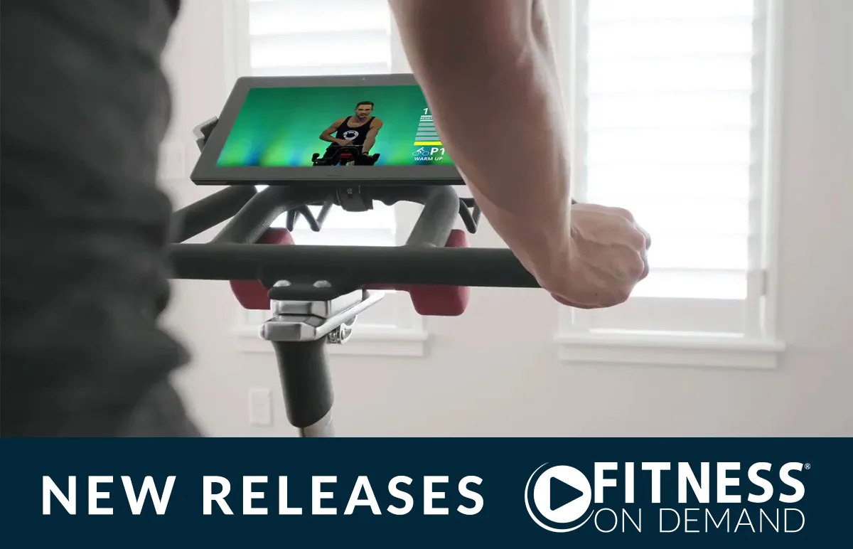 Person riding a stationary bike indoors while watching a virtual fitness class on a screen; text below reads “NEW RELEASES FITNESS ON DEMAND.”.