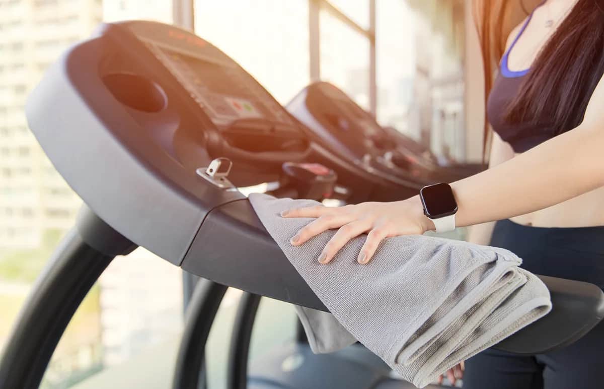 A woman wearing activewear and a smart watch wipes down a treadmill with a gray towel in a bright gym with large windows. Only her arm and upper body are visible.