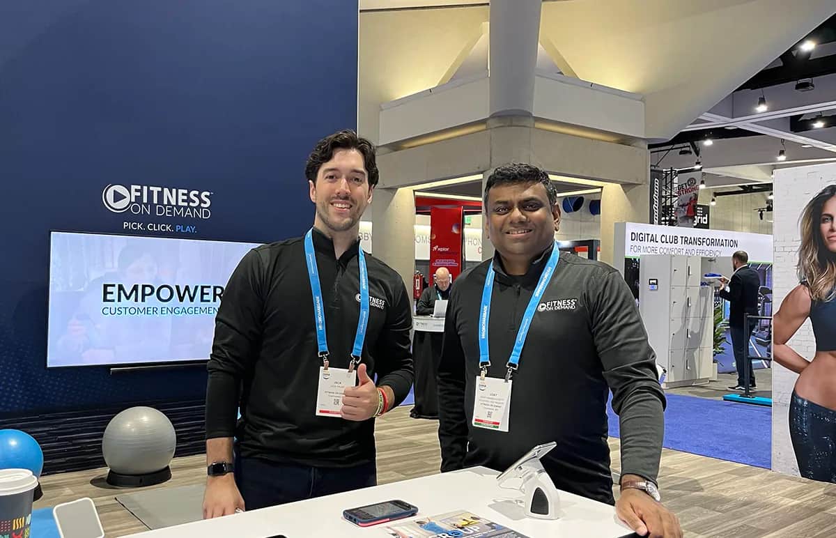 Two men wearing black Fitness On Demand shirts stand smiling at a booth during a convention. A screen behind them displays the company’s logo and the words “Empower Customer Engagement.” Conference badges hang from their necks.