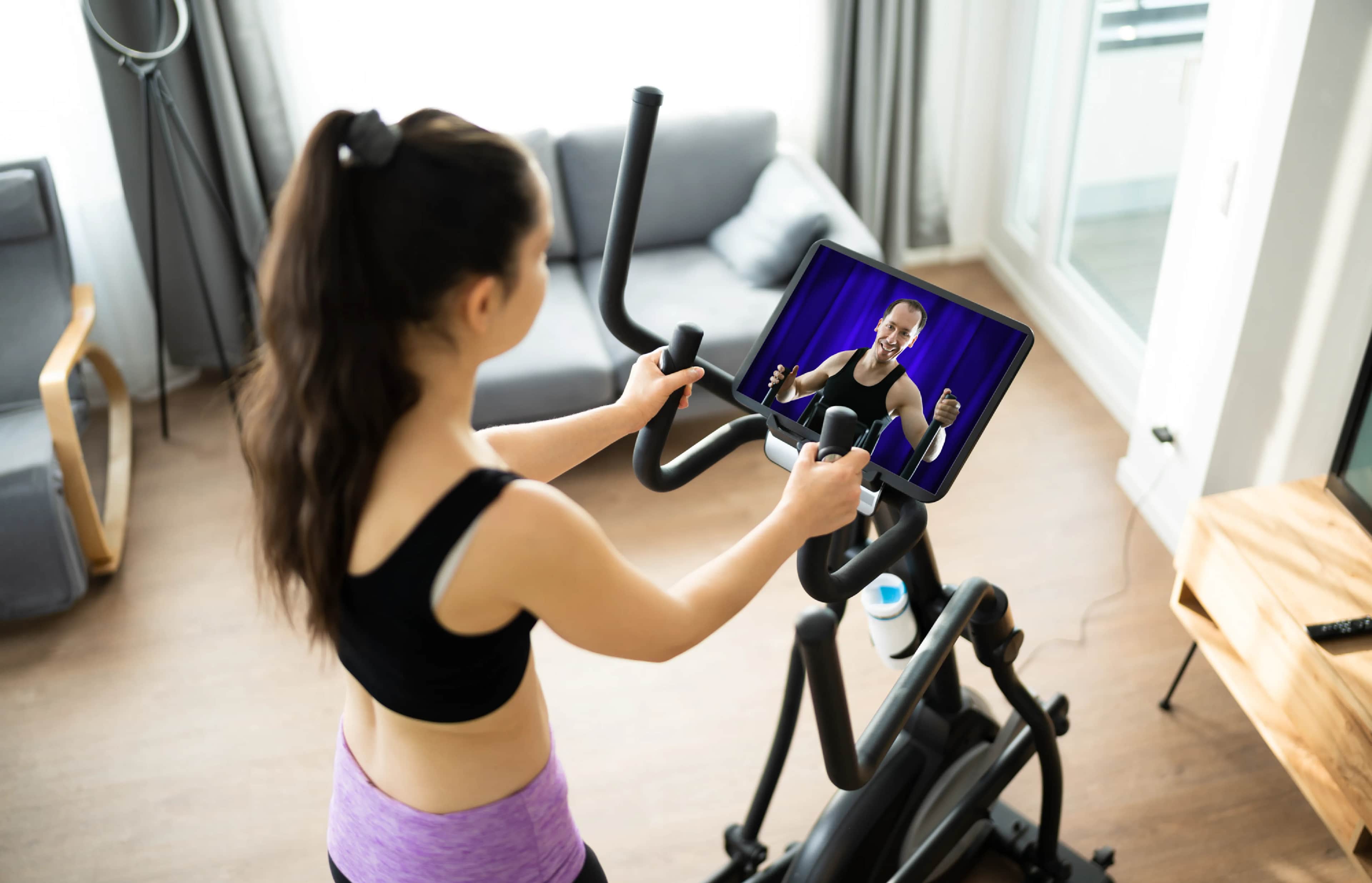Woman exercising on an elliptical machine at home while watching a virtual fitness instructor on a screen in front of her, with a sofa and a window in the background.