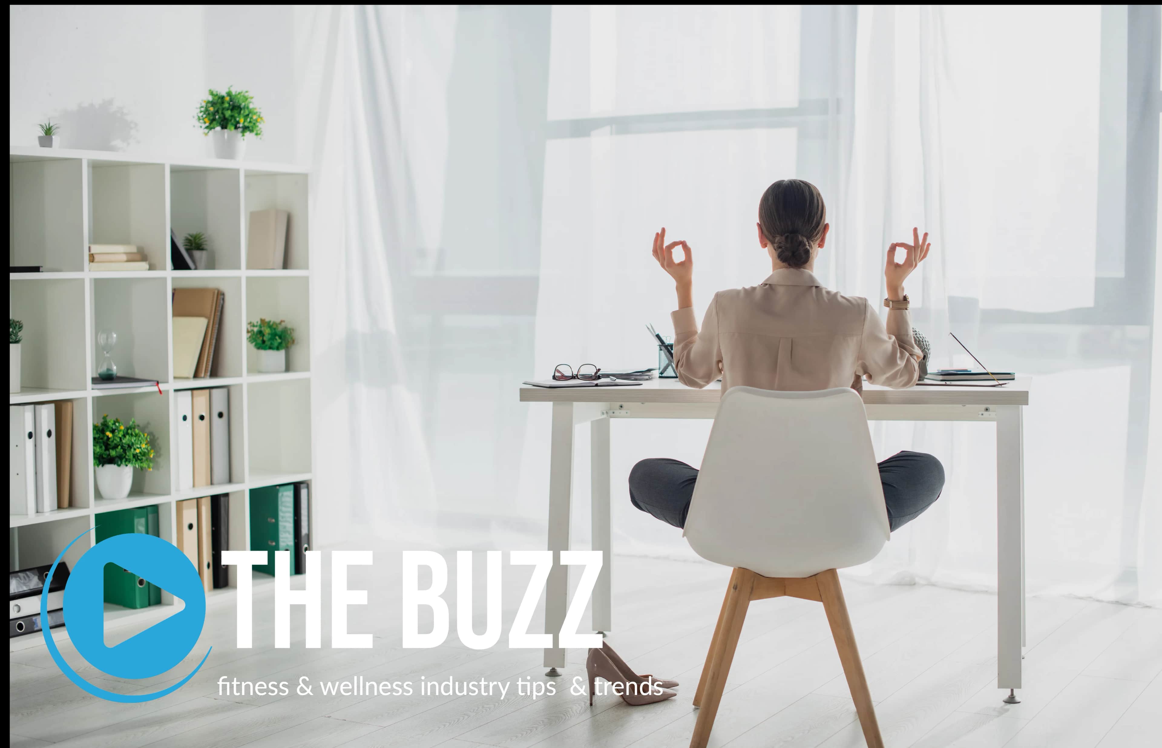 A person sits at a desk in a bright office, facing away, meditating with hands in a yoga pose. The text reads THE BUZZ: fitness & wellness industry tips & trends with a play button logo.