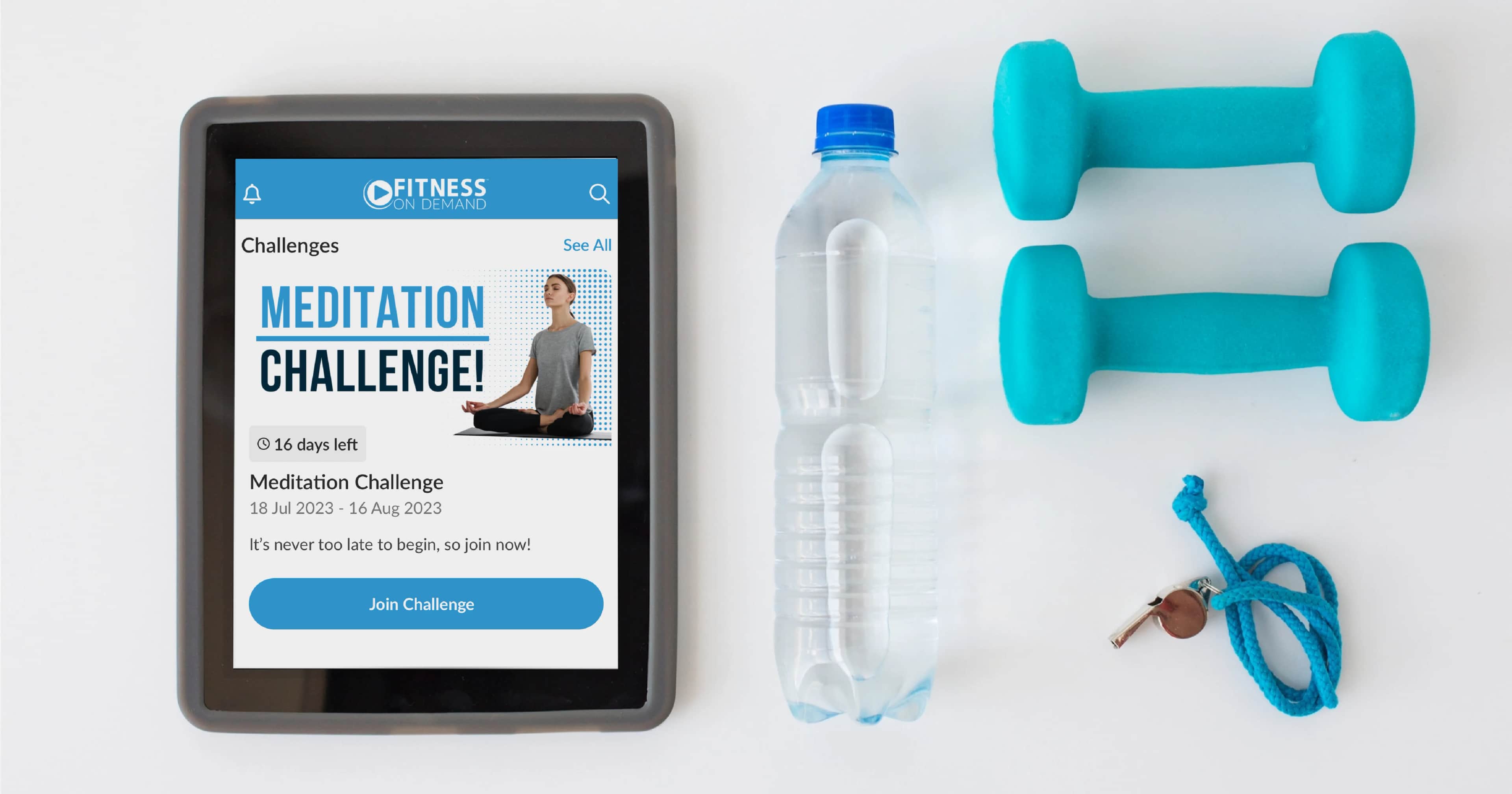 A tablet displaying a Meditation Challenge fitness app screen sits on a white surface next to a water bottle, two blue dumbbells, and a blue jump rope with wooden handles.