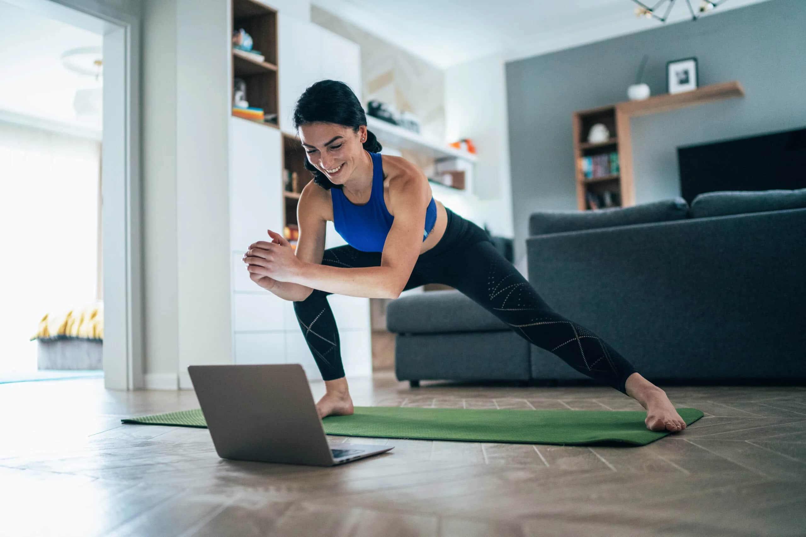 A woman in activewear stretches on a yoga mat in her living room while following a workout on a laptop. She is smiling and appears engaged in her exercise routine.