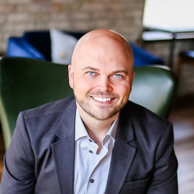 A smiling bald man with a short beard, wearing a gray blazer and light-colored shirt, sits in a modern, softly lit room with a green chair and blurred background.