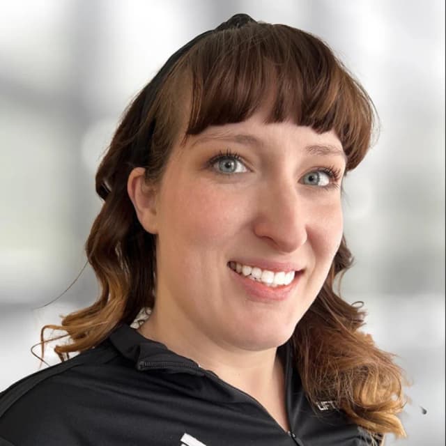 A woman with wavy brown hair and bangs, wearing a black zip-up top, smiles at the camera. The background is blurred and bright.