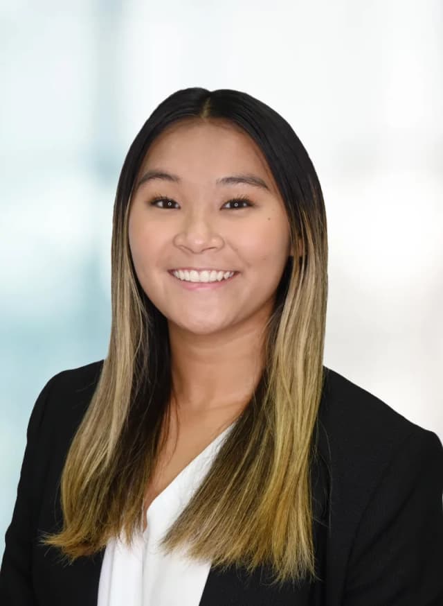 A woman with long, straight dark hair and blonde highlights smiles at the camera. She is wearing a black blazer over a white top, with a blurred light background.
