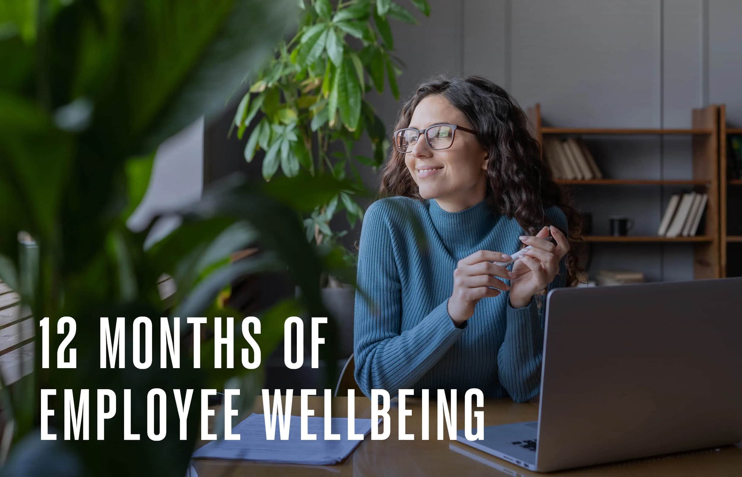 A smiling woman in glasses sits at a desk with a laptop, notebooks, and pen, looking out the window. Green plants and shelves are in the background. Text reads, “12 Months of Employee Wellbeing.”.