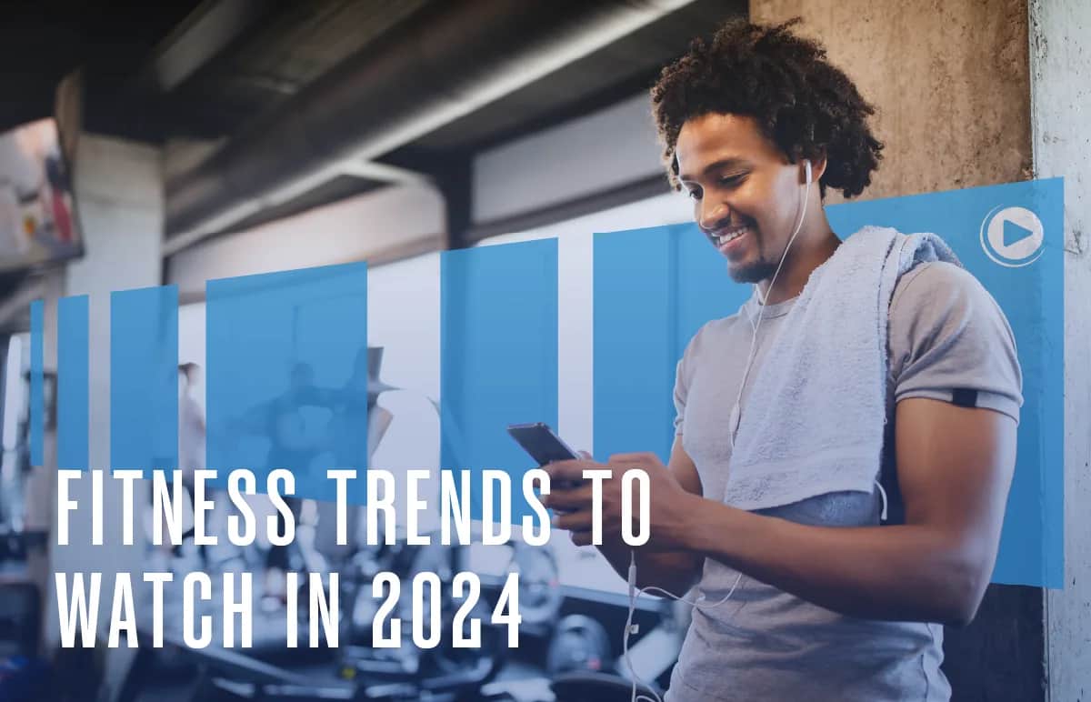 A smiling man with a towel around his neck listens to earphones and looks at his phone in a gym. Text reads: Fitness Trends to Watch in 2024.