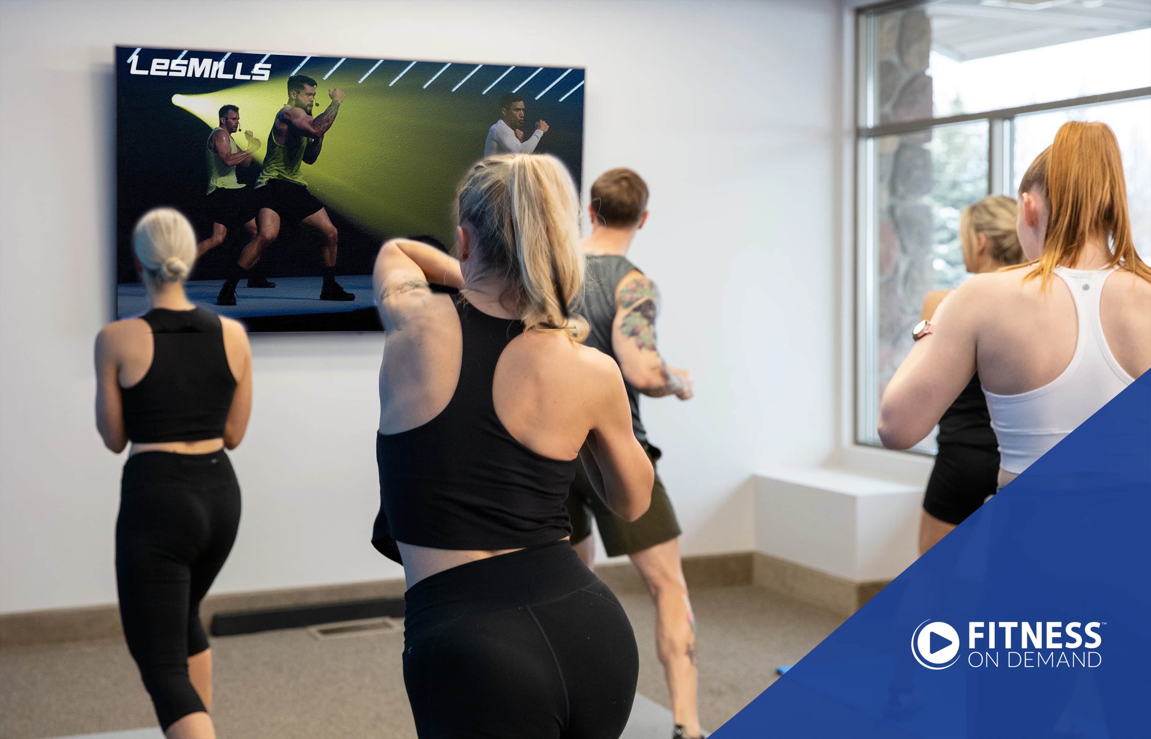 A group of people in athletic wear follow a Les Mills workout video on a screen in a bright fitness studio, with a Fitness On Demand logo in the bottom right corner.