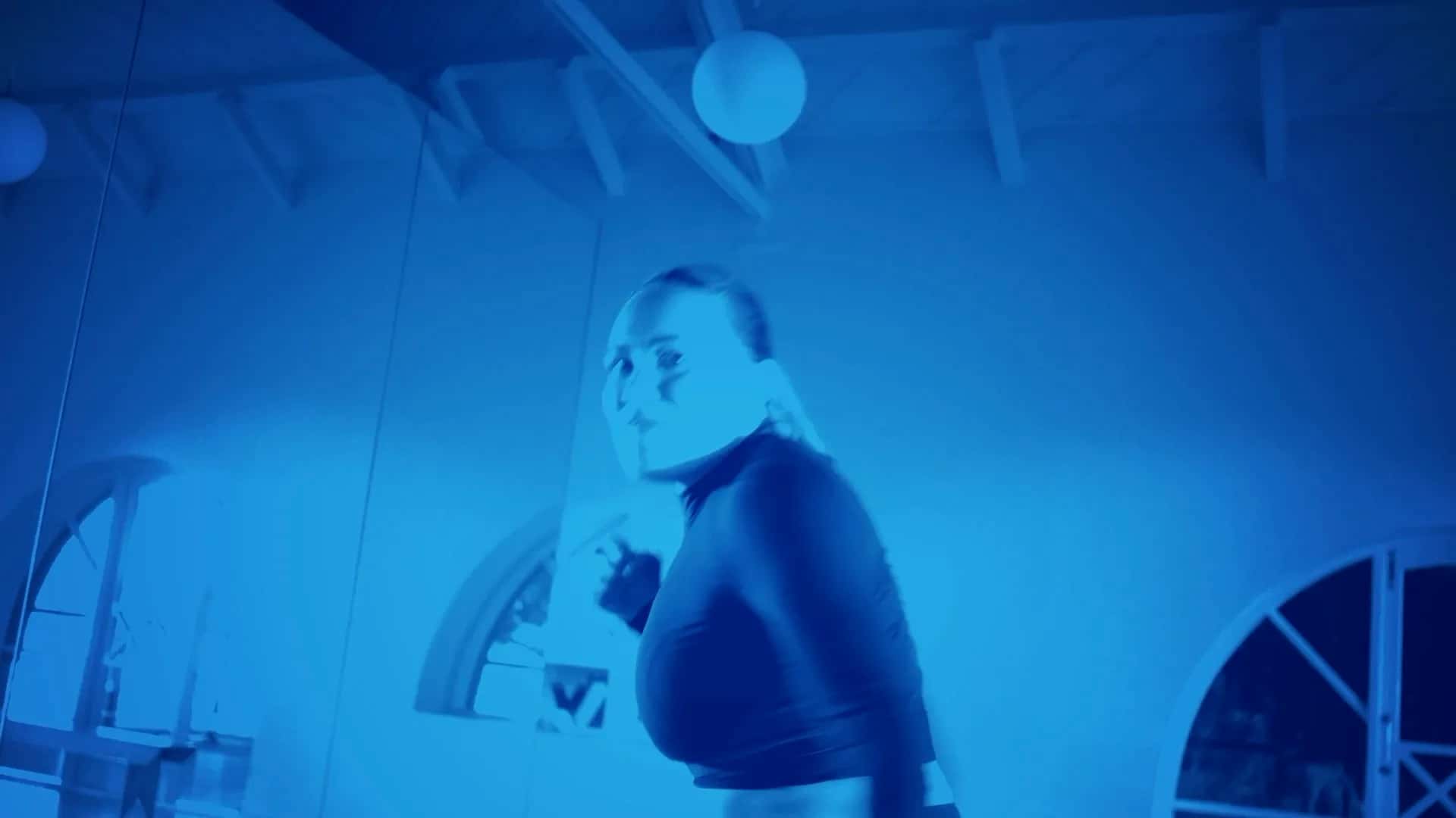 A person wearing a long-sleeve crop top looks over their shoulder in a room with arched windows and exposed beams, illuminated by bright blue lighting.