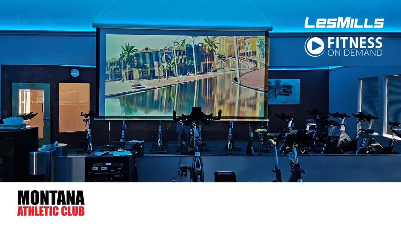 A gym spin room with several stationary bikes faces a large screen displaying a scenic cityscape. Blue lighting fills the room. Logos for LesMills, Fitness On Demand, and Montana Athletic Club are visible.