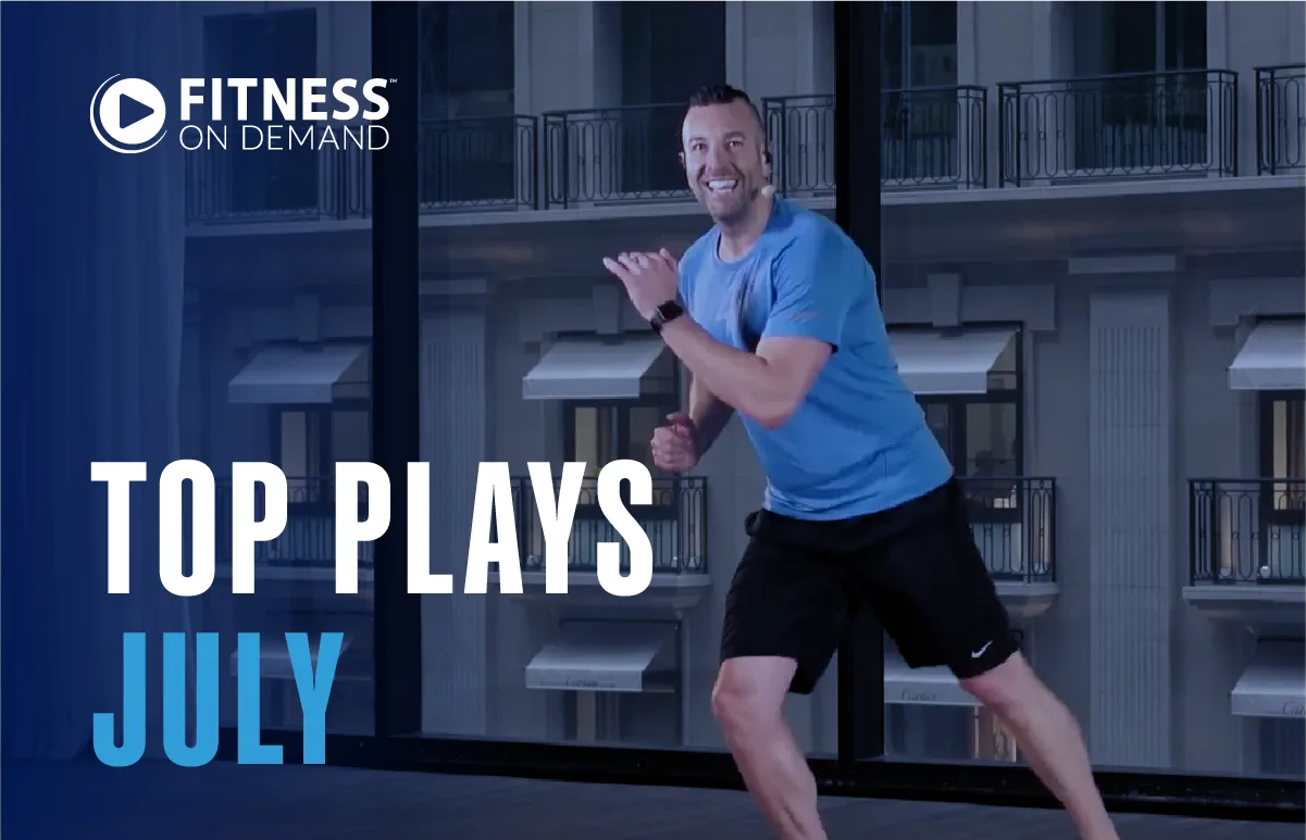 A smiling man in a blue shirt and black shorts exercises indoors. Text reads Fitness On Demand and Top Plays July in front of city building windows.