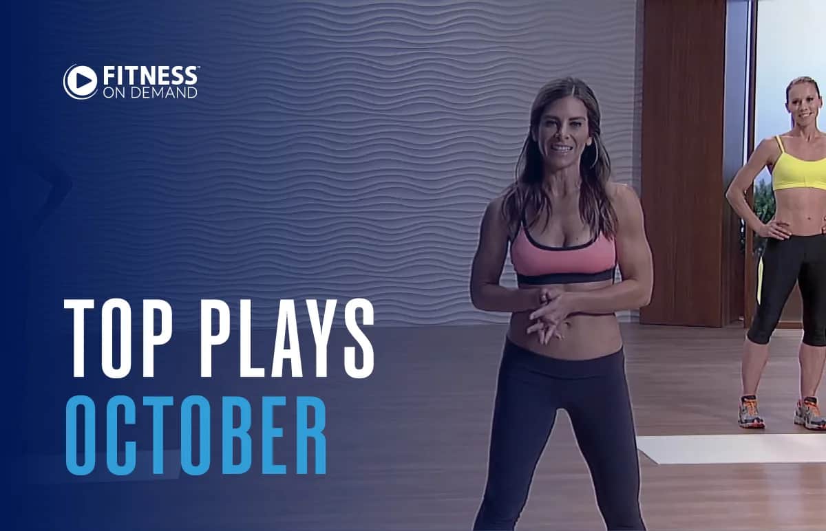 Two women in athletic wear stand in a fitness studio. The text reads Fitness On Demand. Top Plays October. The background features a blue wavy wall design and a wooden floor.