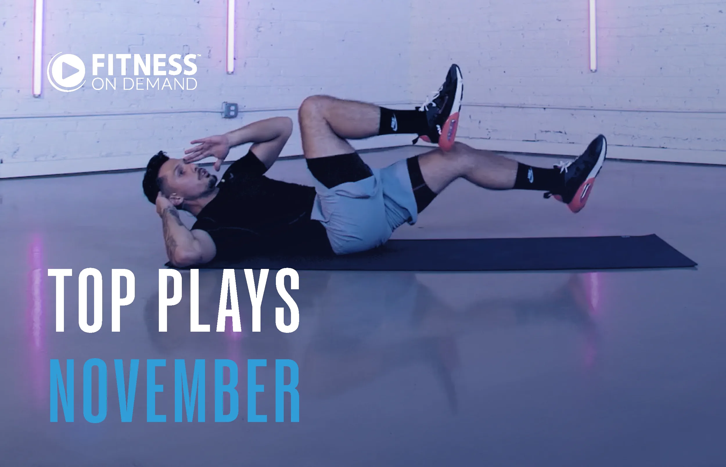 A man exercises on a yoga mat, doing bicycle crunches in a studio with a white brick wall and neon lights. Text reads Top Plays November and Fitness On Demand in the corner.