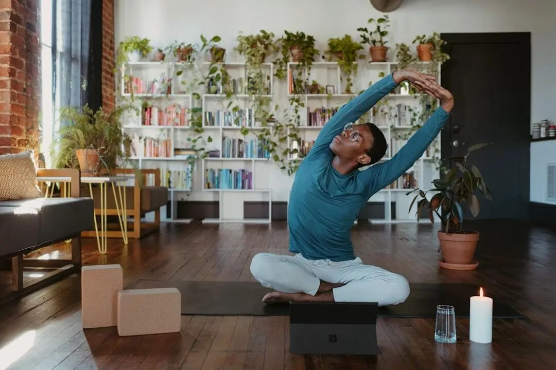 A person sits cross-legged on a yoga mat in a cozy living room, stretching with arms overhead. Nearby are yoga blocks, a candle, a plant, and a laptop. Bookshelves and plants fill the background.