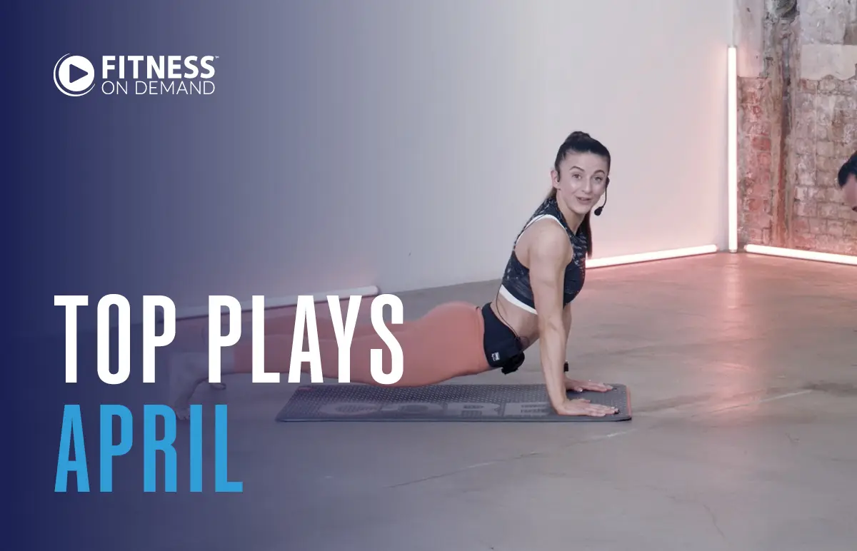 A woman in workout clothes does an upward-facing dog yoga pose on a mat in a studio. Text reads: Fitness On Demand. Top Plays April. The background is minimalist with exposed brick and soft lighting.
