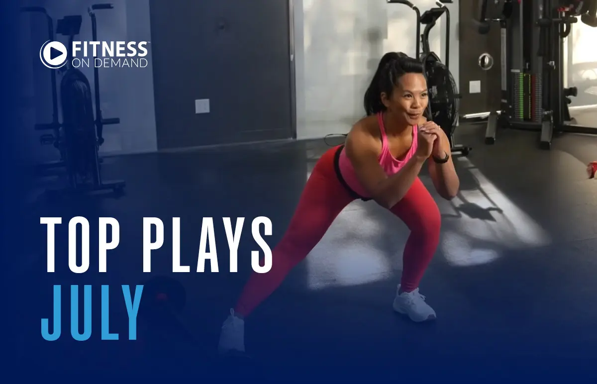 A woman in athletic wear performs a side lunge in a gym. Gym equipment and a large mirror are visible in the background. Text reads Fitness On Demand Top Plays July.