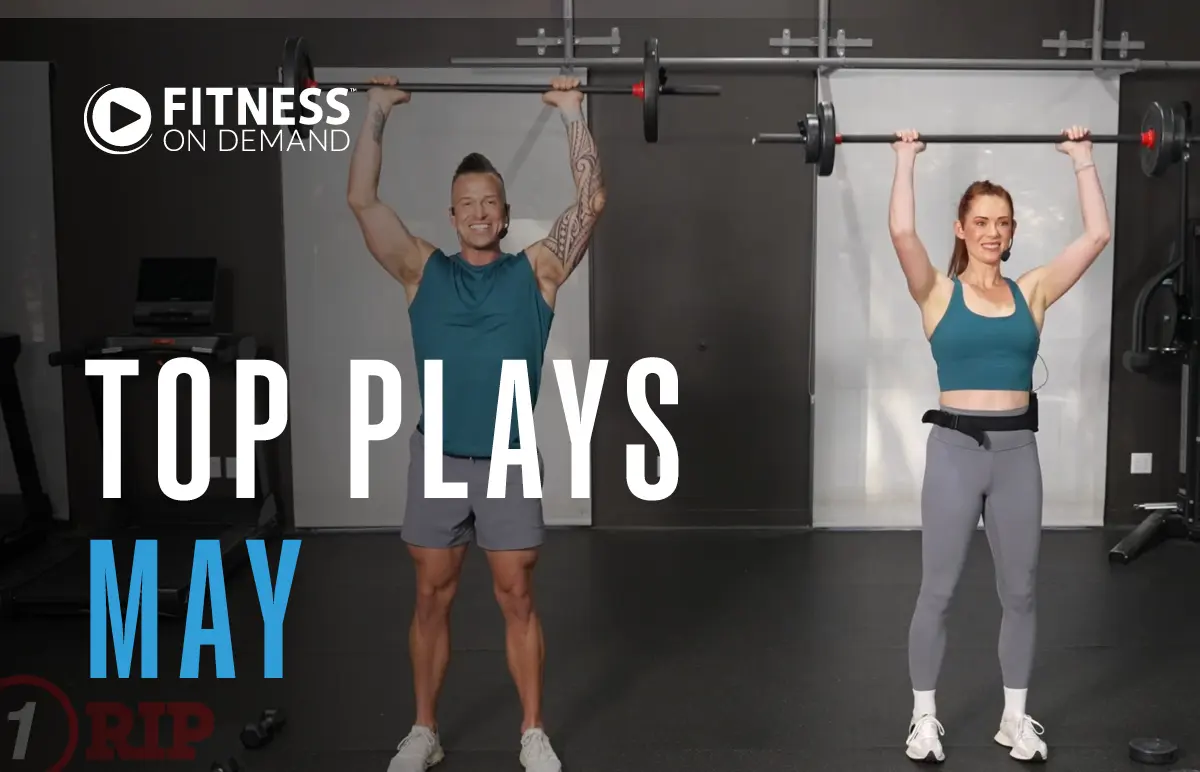 Two people in athletic wear lift barbells overhead in a gym. Text reads: Fitness On Demand Top Plays May. The background shows gym equipment and a dark wall.