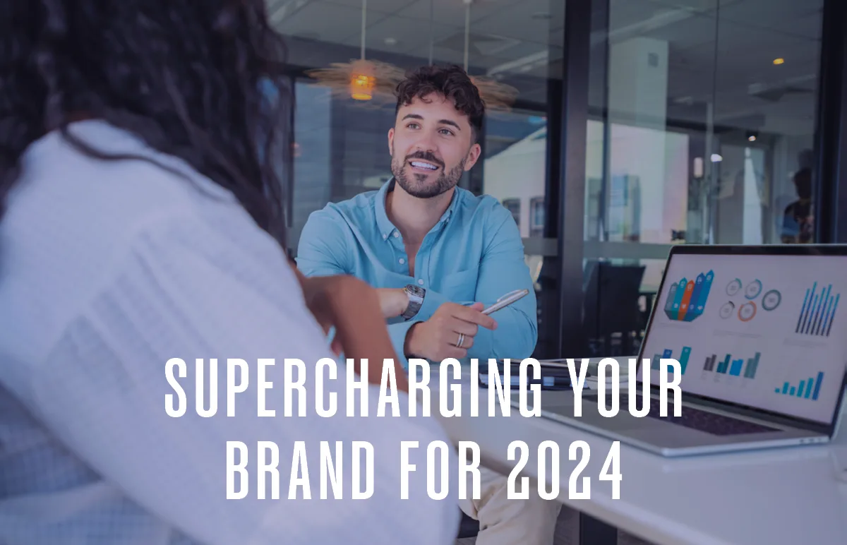 Two people sit across from each other at a desk in a modern office, discussing data shown on a laptop. Text overlay reads, Supercharging Your Brand for 2024.