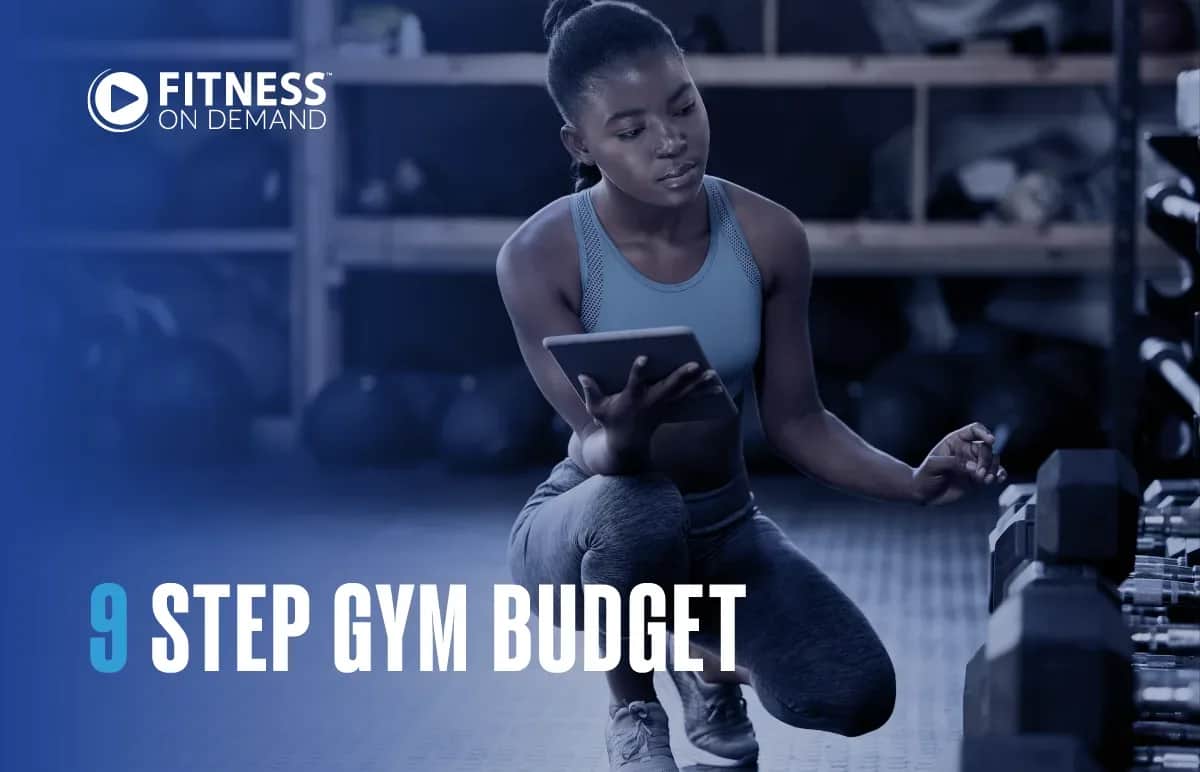 A woman in athletic wear kneels by a rack of dumbbells in a gym, holding a tablet. The text reads Fitness On Demand: 9 Step Gym Budget. The gym background includes weights and shelves.